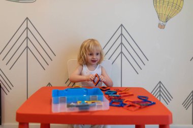 Bir çocuk eğlenceli duvar tasarımlarıyla dekore edilmiş, parlak kırmızı bir masanın üzerindeki renkli yapı taşlarını kullanarak şekiller oluşturmaya odaklanır..