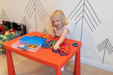 Genç bir çocuk neşeli bir şekilde çeşitli renkli geometrik şekilleri keşfeder, onları doğal ışıkla dolu neşeli bir oyun odasında canlı kırmızı bir masaya dizer ve düzenler..