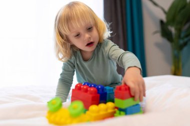 Genç bir çocuk canlı yapı taşlarıyla coşkuyla oynar, onları dikkatlice yığar ve yumuşak bir yatakta parlak, davetkar bir odada çeşitli yapılar oluşturur..