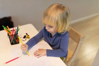 Genç bir çocuk kapalı bir ortamda renkli kalemler kullanarak bir masada çizim yapar. Öğleden sonra yaratıcılığa ve oyunculuğa odaklanacağız..