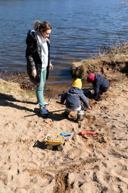 İki çocuk, açık ve güneşli bir günde bir yetişkin onları izlerken, bir nehir kıyısının yakınındaki kumda inşaat ve oyun oynamakla meşgul. Huzurlu ve keyifli bir ortam..