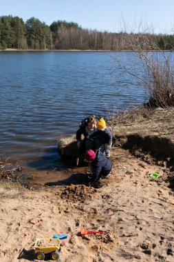 Üç çocuk göl kıyısında güneşli bir günün tadını çıkarırken kumu kazarak eğlenceli aktivitelere girişiyor. Oyuncakları etrafı keşfederken etrafa saçılmış.