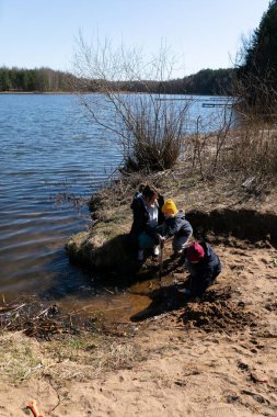 Üç çocuk neşeyle bir nehir kenarında oynuyorlar, kumu kazıyorlar ve sığ suları keşfediyorlar. Güneşli gün, doğal çevrenin güzelliğini arttırır..