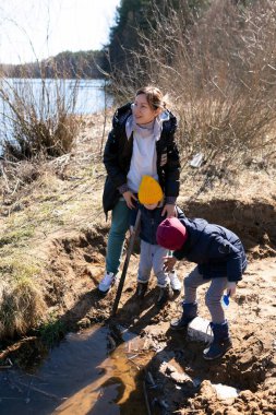 Bir kadın ve iki çocuk, güneşli bir bahar öğleden sonrası çevresel bir çabanın parçası olarak nehir kenarında birlikte çalışarak çöp toplarlar. Su yakınlarda parlıyor, manzarayı güzelleştiriyor..