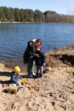 Bir ebeveyn bir çocuğu kucaklarken diğeri gölün kenarında oyuncaklarla kumlarda oynuyor. Sahne ışıl ışıl ve neşeli, aile bağının bir anını yakalıyor..