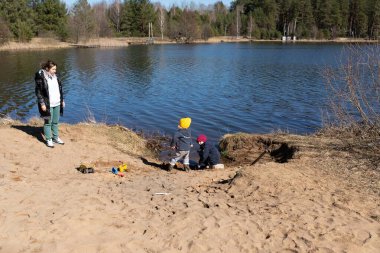 İki çocuk açık bir günde göl kenarında kumu kazarken bir yetişkin kısa mesafeden izliyor. Çevreleyen ağaçlar ve sakin su huzurlu atmosferi güçlendirir..