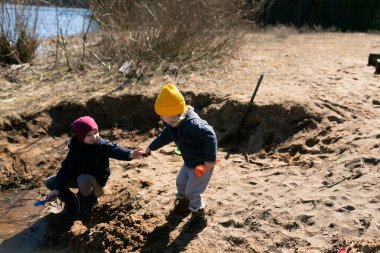 Güneşli bir günde kumlu bir nehir kıyısında oynayan iki çocuk. Bir çocuk kumda otururken, diğeri elini uzatıyor, ikisi de çevrelerini eğlenceli bir keşfe dalmış..