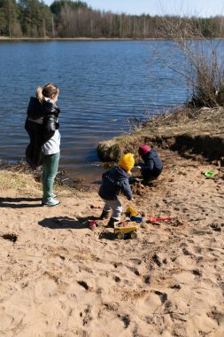 İki çocuk, bir gölün kumlu kıyısı boyunca oyuncakları kazarken ve kullanırken, bir yetişkin, güneşli bir bahar gününde yakınlarda gezinirken, eğlenceli aktivitelere girişir..