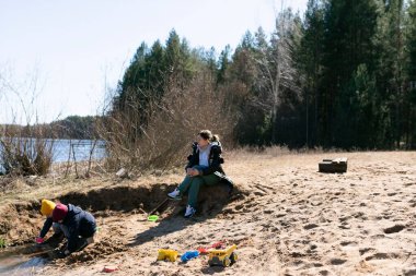İki çocuk bir nehrin yanındaki kumda renkli oyuncaklarla kazıyor ve inşa ediyor. Bir yetişkin kıyıda dinleniyor, ılık bahar havasının ve doğanın tadını çıkarıyor..