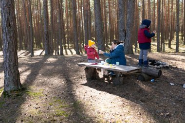 Bir çocuk ve bir yetişkin ormandaki ahşap bir masada abur cubur yiyorlar. Uzun ağaçlar mesafeye kadar uzanıyor, yerde uzun gölgeler bırakıyor..