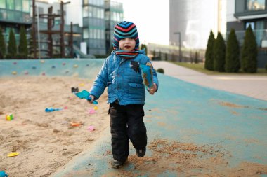 Sıcak giyimli küçük bir çocuk kumlu bir oyun alanında elinde oyuncaklar ve keşifler tutarak yürüyor. Arka planda yeşil ağaçlar ve çağdaş binalar görünür..