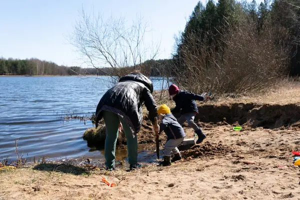 İki genç çocuk sakin bir gölün kenarındaki kumu kazarken, bir yetişkin ormanlık alan yakınlarındaki açık gökyüzünün altında neşeli aktivitelerini denetliyor..