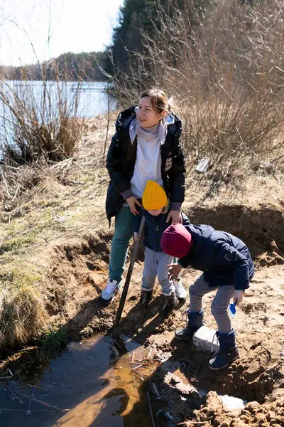 Bir kadın ve iki çocuk, güneşli bir bahar öğleden sonrası çevresel bir çabanın parçası olarak nehir kenarında birlikte çalışarak çöp toplarlar. Su yakınlarda parlıyor, manzarayı güzelleştiriyor..