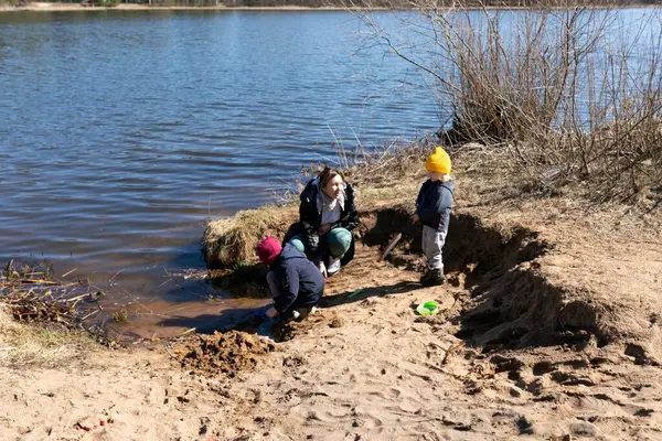 İki çocuk göl kıyısındaki kumda oynamaktan zevk alırken, yakınlarda bir yetişkin güneşli bir günde dinlendirici bir atmosfer sağlıyor..