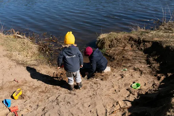 İki çocuk nehir kenarında yaratıcı oyunlar oynuyor, kumu kazıyor ve yapılara şekil veriyor. Parlak renkli giysiler, sıcak bir öğleden sonra güneşlenmelerinin altını çizer..