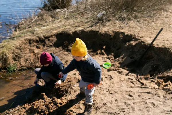 Nehir kenarında güneşli bir günün tadını çıkaran iki çocuk, anne babaları onları izlerken kumda oynayıp deniz kenarını kazıyorlar. Neşeli atmosfer dışarıda eğleniyor..