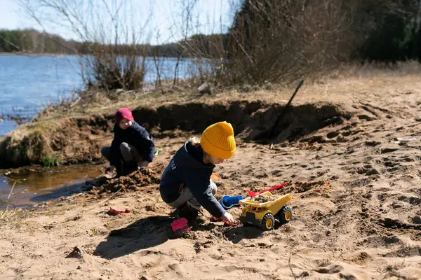 İki çocuk güneşli bir bahar gününde nehir kıyısında eğleniyor. Renkli kum ve oyuncak kamyonlarla yaratıcı bir oyun oynayarak neşeli bir atmosfer yaratıyorlar..