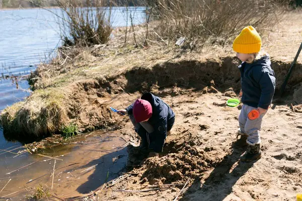 İki çocuk bir nehrin kenarında oynuyor, biri kumda kazarken diğeri küçük bir oyuncak tutuyor. Güneşli hava onların neşeli atmosferine ekleniyor, tipik bir ilkbahar günü gibi..
