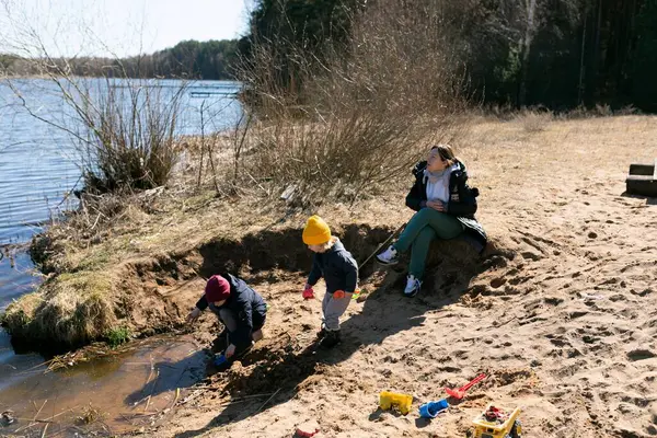 İki çocuk gölün kenarındaki kumu kazarken, bir yetişkin ağaçlarla çevrili güneşli ve sakin bir ortamda dinleniyor..