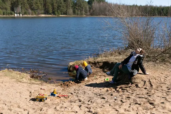 Sıcak bir günde, çocuklar sakin bir gölün kenarında kumlarda oynuyorlar, yapılar inşa ediyorlar. Yetişkin biri günışığının ve huzurlu atmosferin tadını çıkararak rahatça oturur..