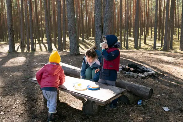 Üç çocuk, berrak mavi gökyüzünün altında uzun çam ağaçlarıyla çevriliyken huzurlu bir ormandaki ahşap bir masanın etrafında atıştırmalıkları paylaşıyorlar..