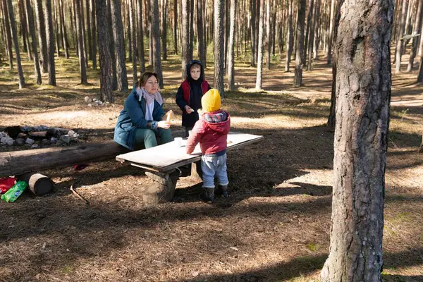 Bir kadın ve bir çocuk, güneşli bir günde neşeli bir atmosfer yaratmak için yakınlarda dikilen başka bir çocuk, sakin bir ormandaki ahşap bir masada oturuyorlar..