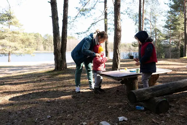 Bir anne çocuğunun sıcak bir ceket giymesine yardım ederken bir başka çocuk yanında duruyor. Göl kenarında bir ormandalar güneşli bir günün tadını çıkarıyorlar piknik masasında birlikte aperatif hazırlıyorlar..