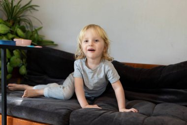 Neşeli bir çocuk rahat bir kanepede rahatlar, gülümser ve eğlenceli bir aktiviteye girişir. Birkaç ev bitkisi oturma odasının sıcacık atmosferine taze bir dokunuş katıyor..