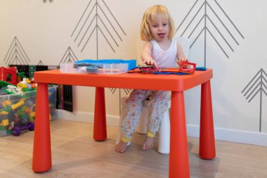 Genç bir çocuk parlak kırmızı bir masada oturmuş basit desenlerle süslenmiş renkli oyuncaklar ve malzemeler düzenlemeye odaklanmış..