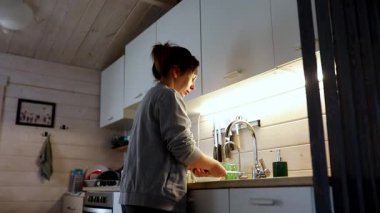 İyi aydınlatılmış bir mutfak lavabosunda duran bir kadın rahat bir atmosferde bulaşıkları yıkıyor. Yumuşak ışık sıcak bir his yaratıyor, görevinin basitliğini vurguluyor..