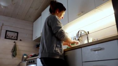 Sıcak, davetkar bir mutfakta bir kadın lavaboda durup bulaşıkları yıkıyor. Yumuşak ışık çalışma alanını aydınlatıyor. Atmosfer, o görevine odaklanırken kırsal bir evde sakin bir akşamı yansıtıyor..