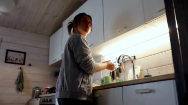 Bir kadın mutfak lavabosunun önünde durup güneş ışığı içeri süzülürken bardağa su döküyor. Sakin atmosfer ve sıcak ışık basit bir mekanda sakin bir sabah rutini oluşturur..