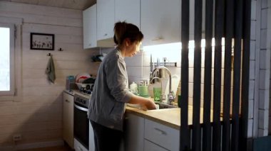 Sıcak ve davetkar bir mutfakta, bir kadın lavaboda bulaşıkları yıkar. Yumuşak ışık çevresini aydınlatır. Uzay, işlevsellik ve konforun bir karışımını sergiliyor.