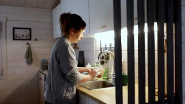 Bir kadın sıcak, davetkar bir mutfakta lavaboda bulaşık yıkamakla meşgul. Yumuşak ışıklandırma, akşam ev işlerini kırsal bir ortamda tamamlarken rahatlatıcı bir atmosfer yaratıyor..