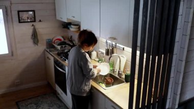 İyi organize olmuş bir mutfakta bulaşık yıkayan bir kadın, hafif ışıkla aydınlatılır. Akşam atmosferi rahatlatıcı ve rahat bir atmosfer yaratıyor..