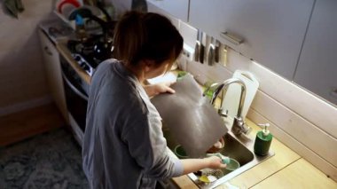 Bir insan, çeşitli mutfak aletleri ve mutfak malzemeleriyle çevrili temiz bir mutfakta bulaşıkları yıkar. Sıcak ışık, akşam yaklaştıkça rahatlatıcı bir atmosfer ekler..