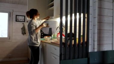 Bir kadın sıcak bir mutfakta akşam yemeğini hazırlamakla meşgul. Yumuşak ışık boşluğu aydınlatırken, o tezgahları ve malzemeleri tezgahın üzerinde düzenler..