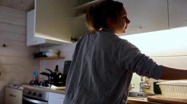 Genç bir kadın akşam yemeğinden sonra bulaşıkları yıkayarak mutfağını topluyor, etrafında sıcak ışıklar ve akşam saatlerinde rahatlatıcı bir atmosfer var..