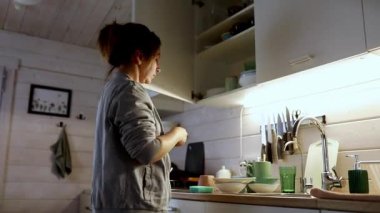 Bir kişi mutfak raflarını düzenliyor ve yemek yapmaya hazırlanırken bulaşıkları yerleştiriyor. Yumuşak ışık, ahşap bir mutfakta sıcak bir atmosfer yaratır..