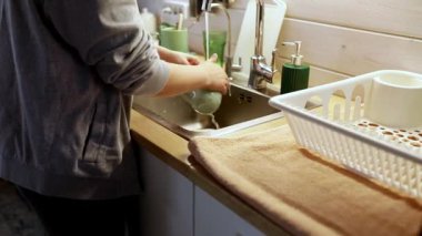 Bir kişi iyi aydınlatılmış bir mutfakta bulaşıkları yıkıyor. Ayarda yeşil aksanlı minimalist dekorlar yer alıyor. Su, fincanı ovalarken akar ve dingin bir atmosfer yaratır..