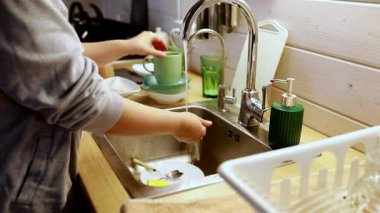 Elleri basit bir mutfakta bulaşık fırçalıyor. Tabaklar ve bardaklar temizlenmek için dizilmişken musluktan su akıyor. Sakin bir atmosfer, akşam yemeği için hazırlanırken boşluğu dolduruyor..