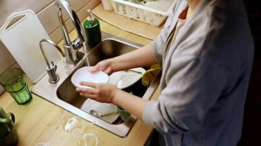 Bir insan mutfak lavabosunun önünde durup akan suyla tabak ve bardakları ovalıyor. Dekor sıcak bir ışık ve rahat bir atmosfer içeriyor. Temizliğe dikkat açıkça görülüyor..