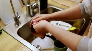 Bir insan mutfak lavabosunda bulaşık yıkıyor, kap kacak ve tabakları sabun ve suyla ovuyor. Mutfakta sade bir atmosfer, mutfak gereçleri ve temiz bir yer var..