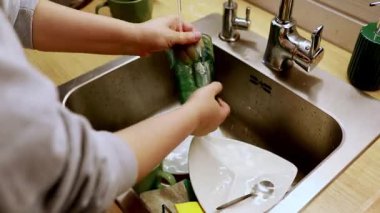 Bir insan iyi aydınlatılmış bir mutfak lavabosunda yeşil bardak ve tabakları temizlemekle meşgul. Onlar kap kacak oyarken musluktan su akar. Tezgah düzenli ve düzenli..