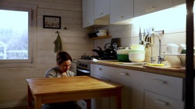 Bir kadın, ahşap bir masaya yaslanırken düşüncelerine dalmış sıcak mutfağının zemininde oturuyor. Yumuşak ışıklandırma, onun yansıma anını geliştiren sakin bir atmosfer yaratıyor..