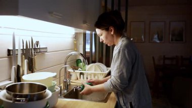 Sıcak ve davetkar bir mutfakta, bir kadın akşam ışığı mekanı hafifçe aydınlatırken lavaboda bulaşıkları yıkamakla meşgul. Atmosfer sakin ve rahat, günlük rutini yansıtıyor..