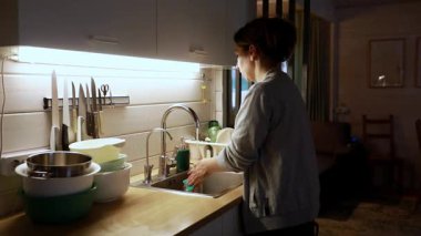 Bir kadın mutfak lavabosunda duruyor, bir yığın bulaşığı, modern mekanı aydınlattığı gibi yıkıyor. Sessiz bir akşam, o etrafı toplarken rahat bir atmosfer yaratıyor..