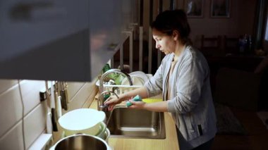 Bir kadın akşamları modern bir mutfakta lavaboda bulaşık yıkıyor. Sıcacık atmosfer, onun toparlanmaya ve düzenlemeye odaklandığını vurguluyor..
