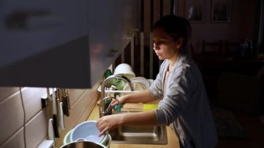 Bir insan yemekten sonra hafif aydınlatılmış bir mutfakta bulaşıkları yıkıyor. Tezgah, sıcak ve davetkar bir atmosferi vurgulayan çeşitli kap ve kaplarla düzenlenmiştir..