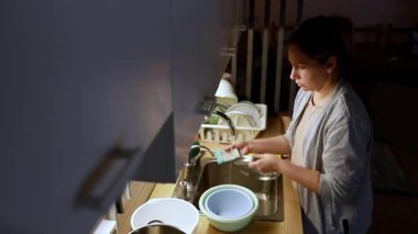 Sıcak bir mutfakta, bir kadın yemekten sonra bulaşıkları yıkar. Lavabodaki eşyaları ovalamaya odaklanıyor. Ortalığı toparlarken sakin bir akşam ortamı yaratıyor..